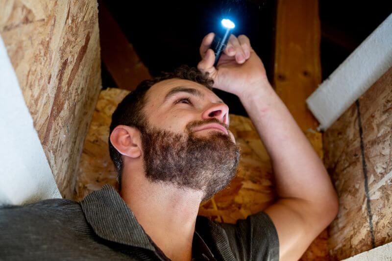 A photo of a man holding a flashlight, looking into an attic in a house.