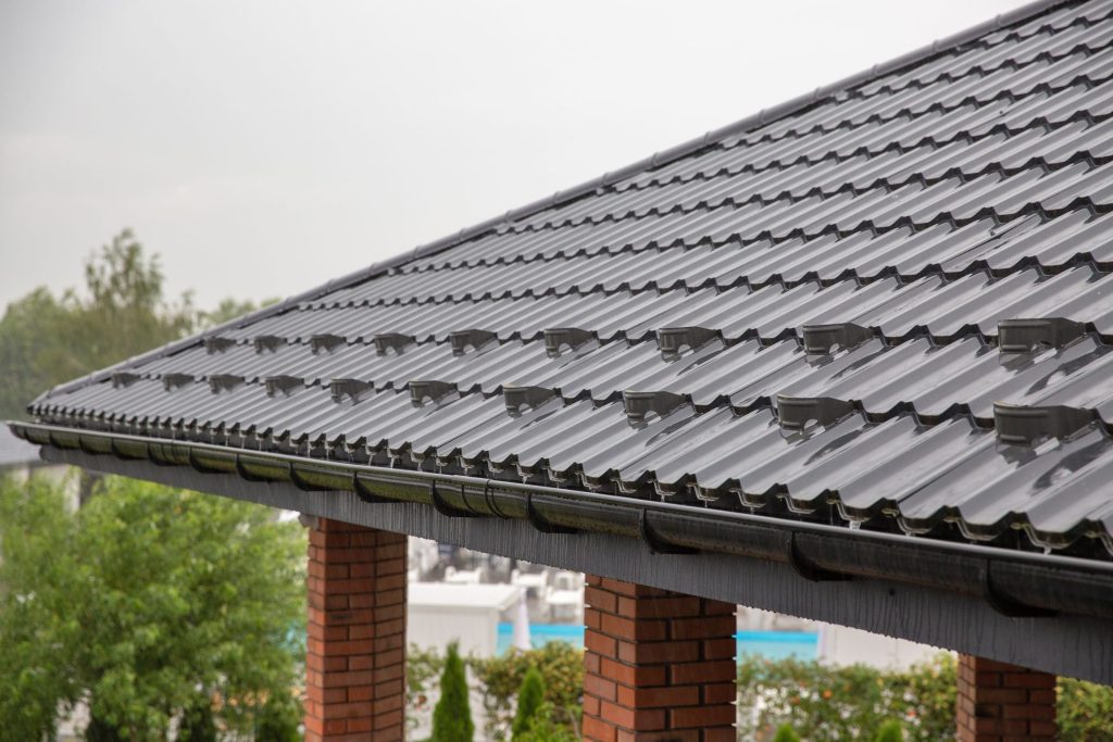 Close-up of dark metal roofing with snow guards installed, supported by brick pillars and surrounded by greenery.