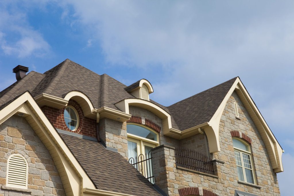 Elegant stone and brick home with high-pitched roof featuring brown impact resistant shingles under a clear blue sky.