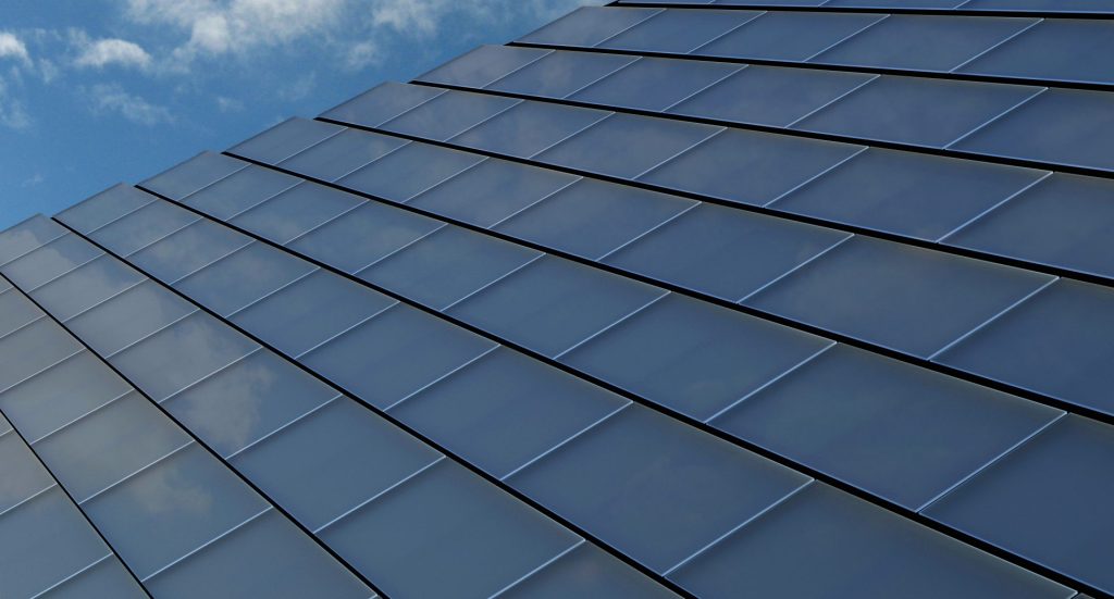 Solar shingles on a modern building facade, angled upward with a clear blue sky and scattered clouds mirrored in the surface.